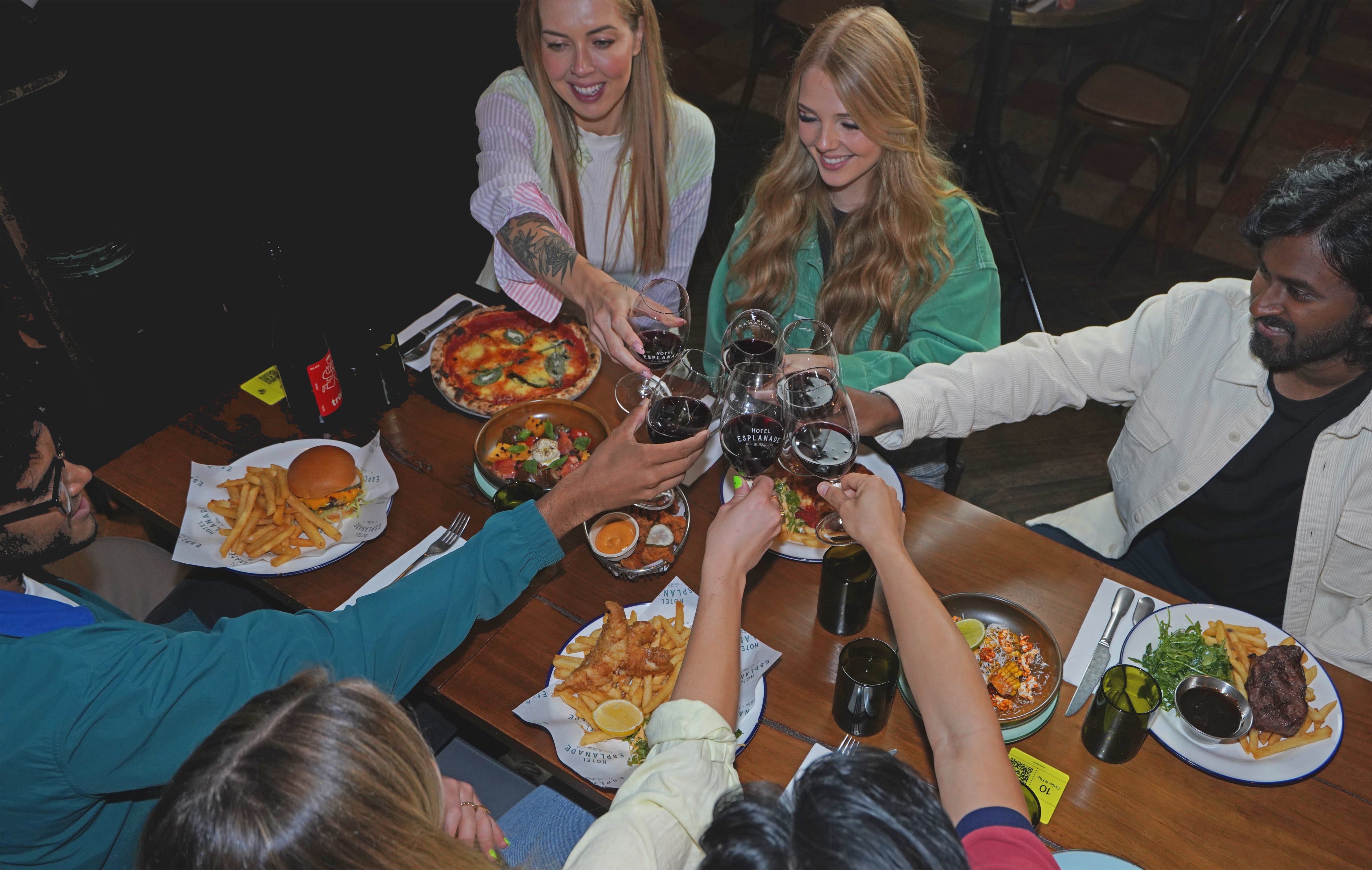 A group of friends sitting around a restaurant table, raising glasses of red wine in a toast. The table is filled with various dishes, including pizza, burgers, fries, tacos, and salads.